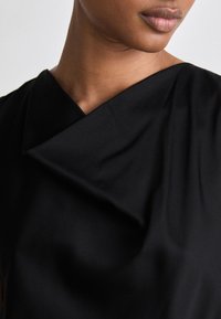 Melna auduma bluze ar drapētu apkakli un gludu tekstūru, ar skaidrām līnijām un maigu spīdumu. Dizains uzsver minimālismu.