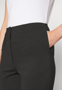 Černé kalhoty s hladkou texturou, vybavené bočními kapsami, s přiléhavým střihem a středním pasem, nošené s bílou košilí.