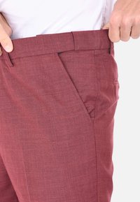 Pantaloni bordeaux realizzati in un tessuto testurizzato, con frontale piatto, passanti per cintura e tasche laterali. La vita è sostenuta da una mano, mostrando la vestibilità.