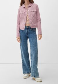 Chaqueta de mezclilla color rosa claro con dos bolsillos en el pecho y botones plateados, combinada con unos vaqueros holgados azules y zapatillas blancas, en una modelo frente a un fondo liso.