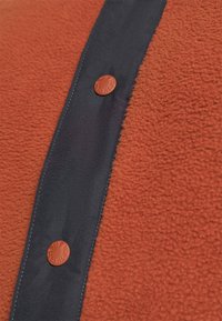 Tessuto in pile di colore arancione bruciato, con una patta in navy e un bottone rotondo marrone con marchio in rilievo, che mette in evidenza una texture morbida.