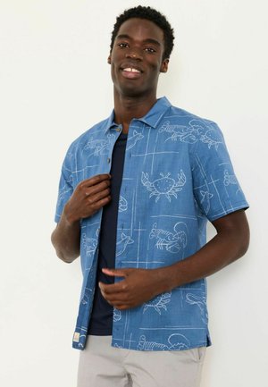 Lächelnder Mann, der ein blaues, kurzärmliges Hemd mit weißen Krabben- und Hummermotiven über einem dunklen T-Shirt trägt.