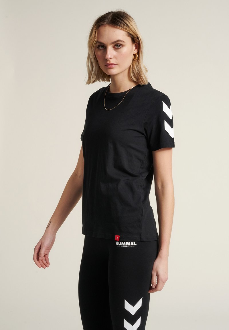 Czarna bawełniana koszulka z krótkim rękawem, z białymi wzorami chevron na ramionach oraz małym czerwonym logo na dole.