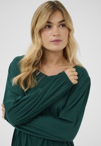 Tamnozeleno dugih rukava s V-izrezom. Mekani, rastezljivi materijal s naboranim manšetama i glatkom teksturom. Silueta koja prati oblik tijela.