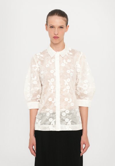 Blouse blanche transparente avec broderie florale, présentant un col classique et des manches bouffantes, boutonnée sur le devant. Contraste de tissu texturé.