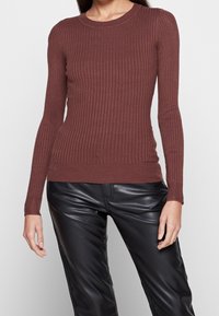 Brązowy prążkowany sweter z długim rękawem, okrągłym dekoltem i dopasowanym krojem, połączony z czarnymi skórzanymi spodniami. Widoczne detale strukturalne materiału.