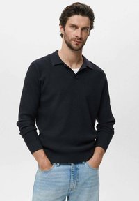 Suéter de punto acanalado azul oscuro con cuello y mangas largas, llevado sobre una camisa blanca. Combinado con jeans azul claro.