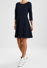 Granatowa dopasowana sukienka z rękawami 3/4, rozkloszowanym dołem i gładkiego materiału. Noszona z białymi botkami za kostkę, ukazując elegancką sylwetkę.