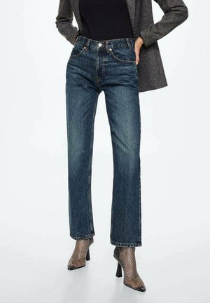 Kobieta w niebieskich dżinsach z wysokim stanem o prostych nogawkach, szarej marynarce, czarnej bluzce i czarnych ażurowych botkach na obcasie, stojąca na białym tle.