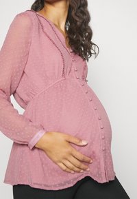 Haut en tissu rose clair à pois avec un design fluide, encolure en V, boutons décoratifs et manches longues transparentes, adapté aux vêtements de maternité.