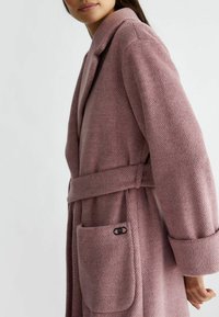 Accappatoio rosa a spina di pesce con colletto, maniche ampie, cintura e tasca laterale con un accento di hardware nero. Tessuto morbido e testurizzato.