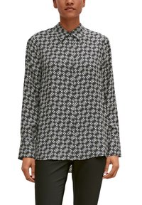 Woman wearing a black and white patterned long-sleeve button-up shirt and black pants, standing against a white background.