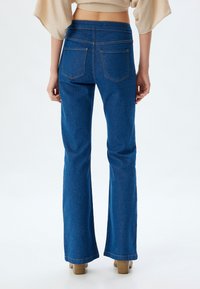 Jeans de mezclilla azul de tiro alto y corte acampanado, con una textura suave, cinco bolsillos y detalles de costura sutiles. El dobladillo se ensancha en la parte inferior.
