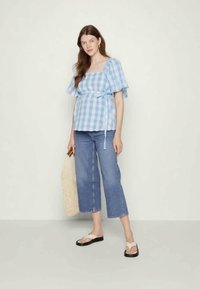 Blusa em padrão xadrez azul e branco com mangas curtas bufantes, atada à cintura, combinada com jeans cropped em azul claro e sandálias claras. Inclui uma bolsa de palha.