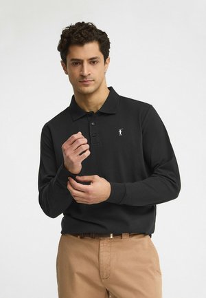 Uomo che sistema il polsino di una polo a maniche lunghe nera con logo ricamato, indossando pantaloni beige su sfondo semplice.