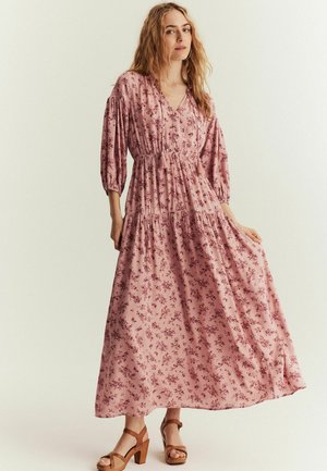 Maxikjole i pink stof med blomsterprint. Har en v-hals, elastisk talje, trekvart pufærmer og flydende kant.