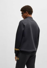 Svart sweatshirt med rund halsringning, långa ärmar och ett guldaccentband på ärmslutet. Slät tygkvalitet och avslappnad passform.