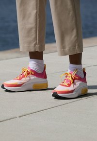 Rosa och vita sneakers med gula accenter, med en överdel i mesh, tjock sula och svart gummitread, vilande på en ljus yta.