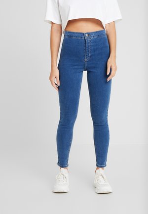 Woman wearing high-waisted blue skinny jeans, white cropped top, and white sneakers, standing against a plain background.