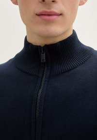 Pulover cu fermoar de culoare bleumarin, cu un guler înalt și textură netedă. Are un închizător central cu fermoar. Vedere detaliată a gulerului.
