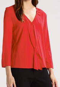 Rød langærmet bluse med V-udskæring, lavet af glat stof, med en draperet frontdesign og subtile plisseringer ved skuldrene.
