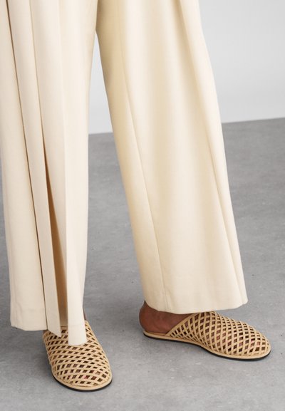 Des chaussures slip-on en cuir tressé beige avec un motif entrecroisé, associées à un pantalon clair à taille haute. Semelle plate et design à dos ouvert.