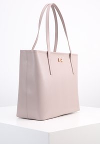 Jasnoróżowa torba tote o zorganizowanej konstrukcji, gładkiej teksturze, z dwoma długimi uchwytami i małym metalowym akcentem w kształcie kokardy z przodu.