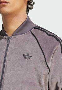 Primer plano de una persona que lleva una chaqueta gris con cremallera, con el logo negro de Adidas y tres rayas negras en el hombro.