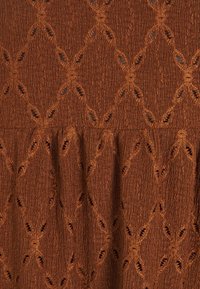 Hnědá texturovaná látka s diamantovými vzory a výřezy, obsahující vertikální linií pro větší hloubku a pohyb.