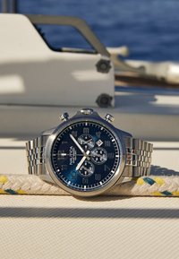 Chronografové hodinky z nerezové oceli s modrým ciferníkem, stříbrnými akcenty, třemi subtitulky a pleteným kovovým náramkem, položené na texturovaném povrchu.