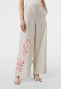 Wijd uitlopende broek in beige stof met een gedurfde rode bladprint aan de linkerkant en een platte tailleband. Draagt bijpassende witte sandalen met bandjes.