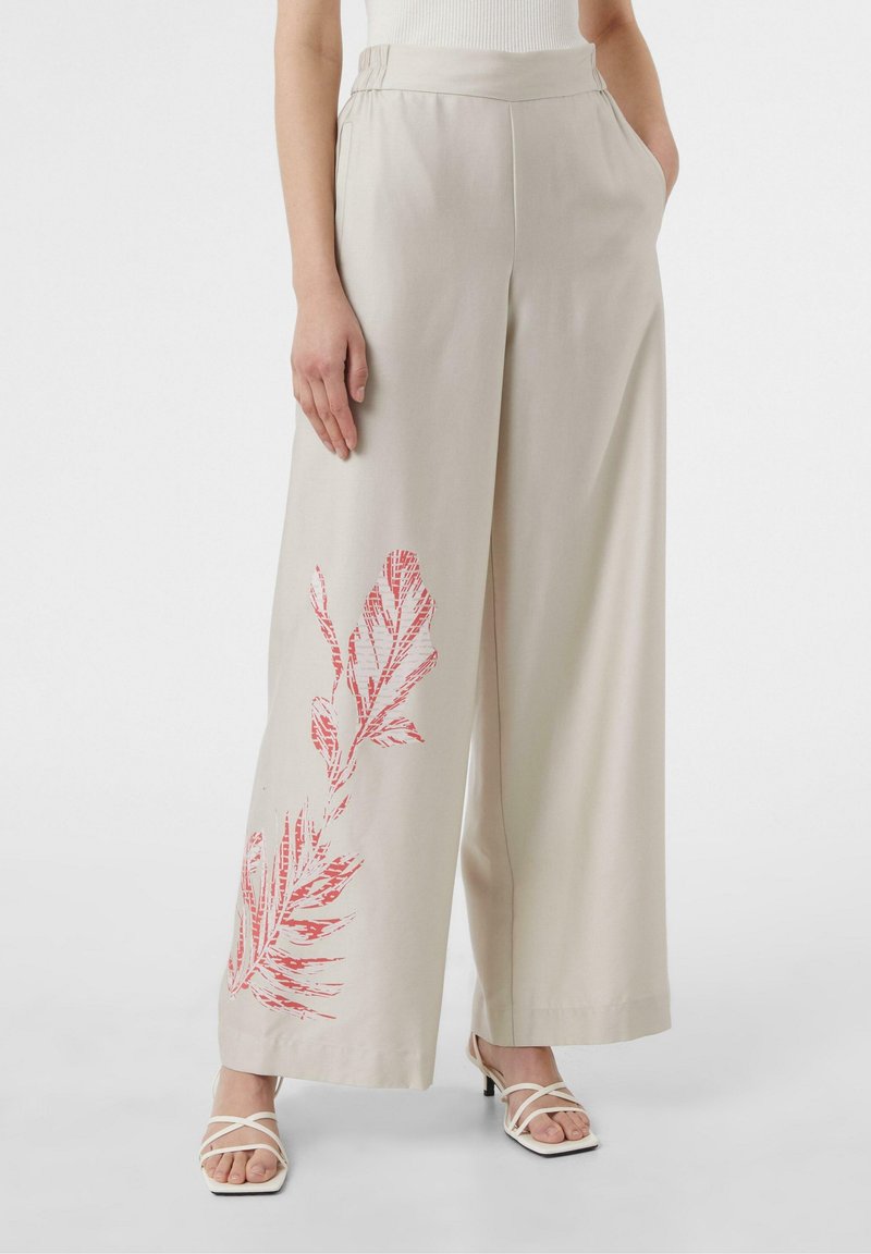Wijd uitlopende broek in beige stof met een gedurfde rode bladprint aan de linkerkant en een platte tailleband. Draagt bijpassende witte sandalen met bandjes.
