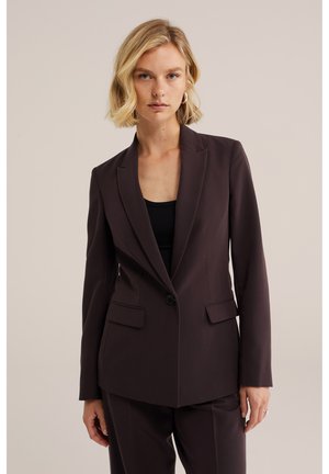 Femme aux cheveux blonds courts portant un blazer marron foncé taillé sur mesure et un pantalon assorti sur un fond uni.