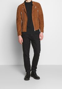Giacca biker in suede marrone con tasche con zip, abbinata a una t-shirt nera e jeans neri. Stivali neri completano l'outfit.