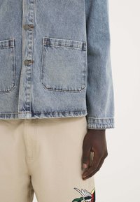 Vaatetus vaaleansininen farkkutakki, jossa on kaksi etutaskua, napitus, ja kontrastiväriset tikit; yhdistetty beigen brodeerattujen housujen kanssa.