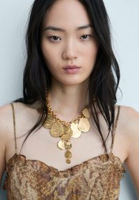Collana dichiarativa in oro con pendenti ovali texturizzati, catena texturizzata, indossata sopra un top marrone a fantasia con bordi arricciati.