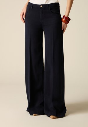 Kobieta w czarnych, szerokich spodniach z wysokim stanem, z przodu zapinanych na guziki i z kieszeniami, w połączeniu z beżowymi, szpiczastymi butami i drewnianymi bransoletkami.