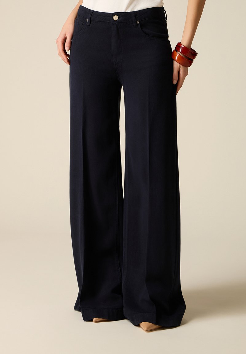 Femeie purtând pantaloni negri cu talie înaltă și croială largă, cu nasturi în față și buzunare, asortați cu pantofi bej cu vârf ascuțit și brățări din lemn.