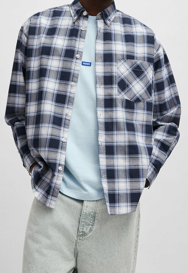 Chemise à carreaux boutonnée en marine, blanc et gris ; elle comporte une poche poitrine, un col et des manches longues portées sur un t-shirt bleu clair.