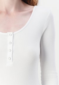 Biała prążkowana bluzka z długim rękawem, z okrągłym dekoltem i pięcioma metalowymi napami z przodu. Gładka tekstura.