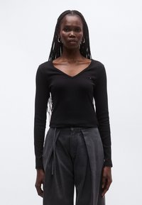 Musta pitkähihainen V-kaula-aukollinen toppi, joka on tehty ribbineuloksesta ja jossa on pieni logo lähellä rintaa, yhdistetty korkeavyötäröisiin harmaisiin housuihin.