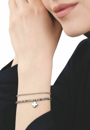 Pols met twee zilveren armbanden, één met kralen en één met een klavertjevier-bedel, dicht bij iemands lippen en kin tegen een witte achtergrond.