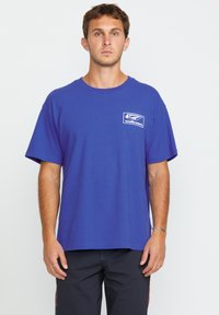 Blauwe katoenen T-shirt met een logo op de borst in het wit. Ronde hals en korte mouwen; ontspannen pasvorm. Glad van textuur, effen kleur zonder patronen.