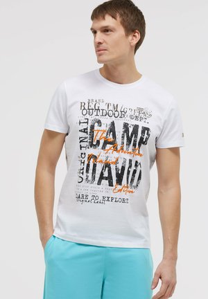 Junger Mann schaut seitlich, trägt ein weißes T-Shirt mit schwarzem und orangem Schriftzug und hellblaue Shorts.