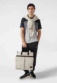 Bolso tote beige con detalles negros, dos bolsillos delanteros y asas resistentes. El modelo lleva una camiseta gris y negra, pantalones de chándal oscuros y zapatillas.