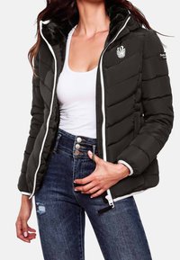 Zwarte pufferjack met een rits aan de voorkant, een capuchon en witte accenten. Heeft een gestikt patroon, zachte furry voering en geborduurd logo op de borst.