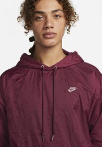 Maroon Nike jakna s kapuco s teksturo mečkanega materiala, opremljena z vrvico za zategovanje kape, preprostim belim logotipom in lahkotno zasnovo.