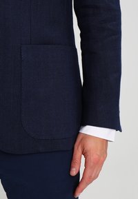 En mans hand som vilar bredvid en marinblå kavaj med synlig framficka och vit skjortärm, mot en neutral bakgrund.