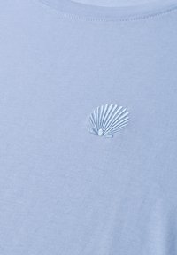 Tissu bleu clair avec une petite coquille blanche brodée en détail près de l'encolure d'un vêtement.