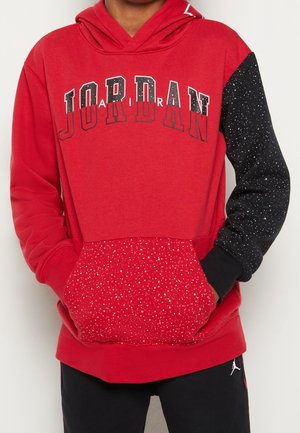 Czerwona bluza z kapturem z czarno-białym nakrapianym prawym rękawem i kieszenią, z napisem „JORDAN” na klatce piersiowej w czarno-białych literach.